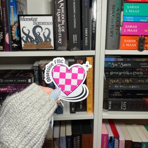 May include: A pink and white checkered heart sticker with a black outline and the text "Obsessive FMC House Bane Brood" in a white circle around the heart.