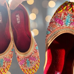 May include: A pair of beige embroidered flat shoes with colorful beads and sequins. The shoes have a red velvet lining and the brand name "HER" is printed on the inside.