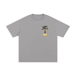 May include: A grey short-sleeved t-shirt featuring a small graphic on the left chest. The graphic displays the text "STOTOK" in black script above a small illustration of a soccer player in a yellow and black kit standing on a yellow base.