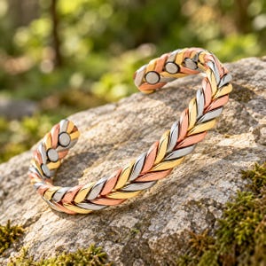 Könnte beinhalten: Ein dreifarbiger Armreif mit geflochtenem Design aus abwechselnden Segmenten in Gold-, Silber- und Kupferoptik. Das offene Armband zeigt an den inneren Enden deutlich sichtbare, kreisförmige magnetische Einsätze.