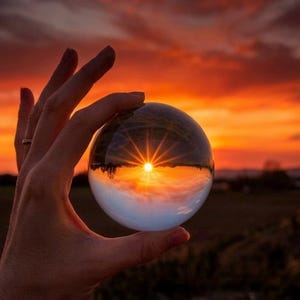 Peut inclure: Une sphère de verre transparente tenue dans une main, reflétant un coucher de soleil vibrant. Le coucher de soleil présente un éclat solaire lumineux et un ciel orange et rouge flamboyant. La sphère capture le paysage et le ciel.