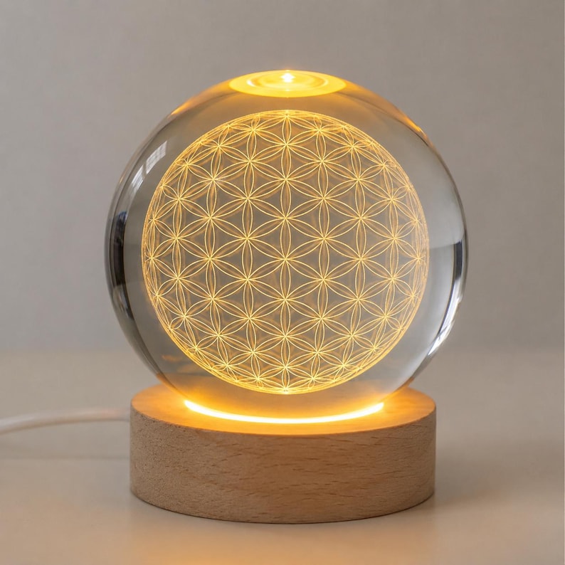May include: A clear spherical glass lamp featuring an internal laser-etched flower of life geometric pattern. The sphere rests on a cylindrical light-colored wooden base that illuminates the glass from below with a warm yellow glow.