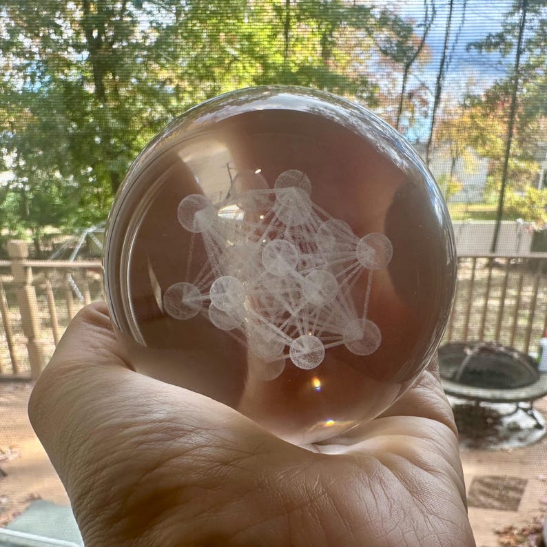 May include: A clear, spherical glass paperweight containing an internal white laser-etched geometric pattern. The design features a complex arrangement of interconnected spheres and lines, creating a three-dimensional structure suspended within the solid glass orb.