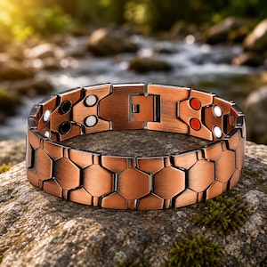 Könnte beinhalten: Ein kupferfarbenes Gliederarmband mit einem sich wiederholenden Sechseckmuster. Die Innenseite des Armbands zeigt kleine kreisförmige Akzente in Weiß, Schwarz und Rot sowie einen sicheren Metallverschlussmechanismus zum Schließen.