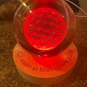 May include: A clear glass sphere containing an internal laser-etched flower of life pattern illuminated by a bright red light. The sphere rests on a circular light-colored wooden base engraved with the text HMH at Metropark 2026.