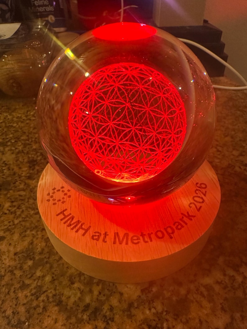 May include: A clear glass sphere containing an internal laser-etched flower of life pattern illuminated by a bright red light. The sphere rests on a circular light-colored wooden base engraved with the text HMH at Metropark 2026.