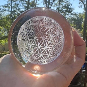 May include: A clear, spherical glass paperweight featuring an intricate, white, laser-etched Flower of Life geometric pattern suspended within the center. The transparent orb reflects light, highlighting the detailed, repeating circular design.