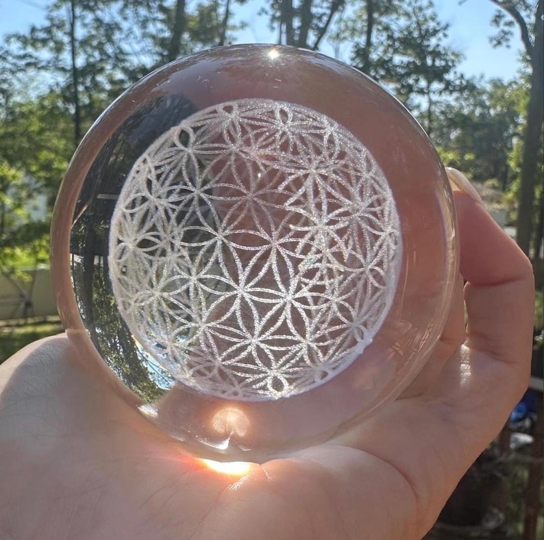 May include: A clear, spherical glass paperweight featuring an intricate, white, laser-etched Flower of Life geometric pattern suspended within the center. The transparent orb reflects light, highlighting the detailed, repeating circular design.