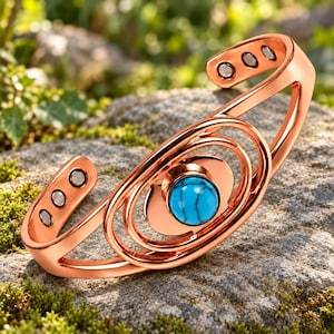 Könnte beinhalten: Ein Kupfer-Armreif mit einem zentralen, kreisförmigen Türkisstein mit dunkler Maserung. Das Armband hat ein offenes Design mit konzentrischen ovalen Formen und drei kleinen, runden Magneten an jedem inneren Ende des Bandes.