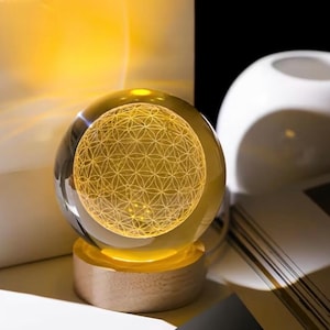 May include: A clear glass sphere resting on a cylindrical light-colored wooden base. The interior of the sphere features a glowing yellow laser-etched geometric pattern of overlapping circles, known as the flower of life, illuminated from below.