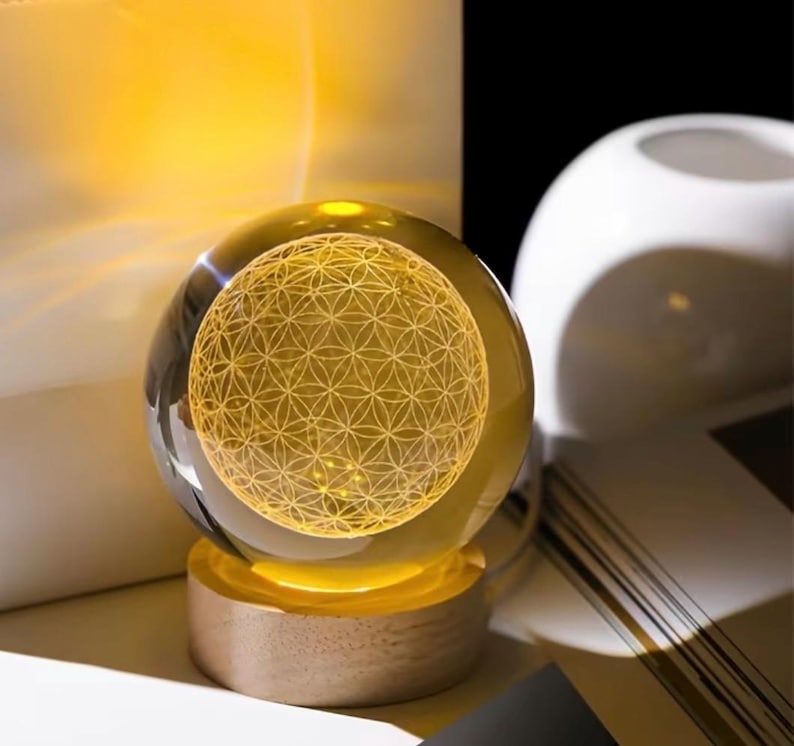 May include: A clear glass sphere resting on a cylindrical light-colored wooden base. The interior of the sphere features a glowing yellow laser-etched geometric pattern of overlapping circles, known as the flower of life, illuminated from below.