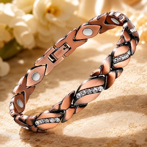 Puede incluir: Una pulsera de eslabones en tono cobre con secciones alternas de eslabones en forma de X con imanes circulares plateados incrustados y eslabones en forma de hoja con cristales transparentes. El cierre tiene grabado el texto copper.