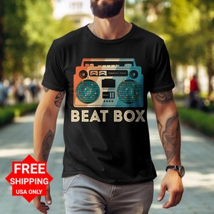 Puede incluir: Una camiseta negra con una ilustración de estilo vintage de una radiocassette con bolas de discoteca a cada lado. El texto "BEAT BOX" está impreso debajo de la radiocassette.