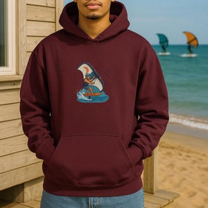 Peut inclure: Un sweat à capuche bordeaux avec un motif représentant une personne faisant de la planche à voile sur une vague. Le motif est de style vintage avec une palette de couleurs bleu, blanc et orange.