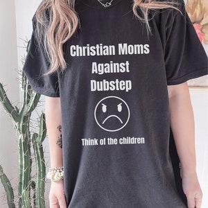 May include: Black t-shirt featuring the text "Christian Moms Against Dubstep" in white, with a sad face graphic and the words "Think of the children." The shirt has a relaxed, oversized style.