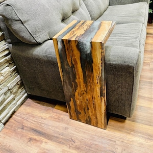 May include: A wooden side table with a live edge design. The table has a natural wood grain pattern with a clear epoxy resin inlay. The rectangular table is designed to fit over a sofa arm. The wood tones are brown and tan.