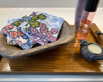 May include: A stack of square fabric facial rounds featuring a floral pattern in shades of pink, blue, and green. The rounds are placed inside a rustic, oval-shaped wooden bowl, which sits on a rectangular wooden tray alongside a bird-shaped candle holder.