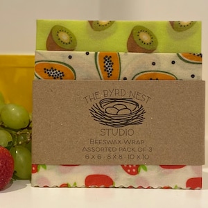 May include: A set of three beeswax food wraps with different fruit designs. The wraps are in a stack, with a kiwi design on top, a papaya design in the middle, and a strawberry design on the bottom. The wraps are packaged in a brown paper envelope with the text "The Byrd Nest Studio Beeswax Wrap Assorted Pack of 3 6 x 6 - 8 x 8 - 10 x 10".