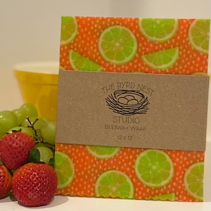 May include: Beeswax wrap with a vibrant orange background and lime slices. The wrap is labeled "THE BYRD NEST STUDIO BEESWAX WRAP 12 x 12". Strawberries, grapes, and a yellow bowl are in the background.