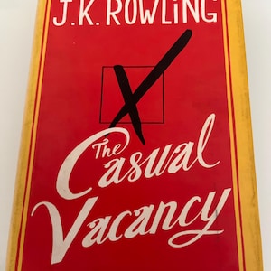 May include: A hardcover book titled "The Casual Vacancy" by J.K. Rowling. The book cover is red with white lettering and a black checkmark inside a square. The book has a yellow border.