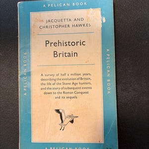 May include: A vintage paperback book titled "Prehistoric Britain" by Jacquetta and Christopher Hawkes. The cover is light blue with a cream-colored central panel. The book features a Pelican Books logo and a drawing of a bird.