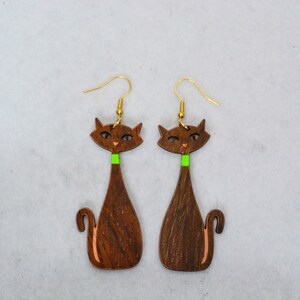 Pode incluir: Um par de brincos de gato de madeira. Os brincos apresentam uma silhueta de gato castanho escuro com uma gola verde e ganchos de orelha dourados. O rosto do gato tem pequenos detalhes laranja. Os brincos s&atilde;o projetados com uma est&eacute;tica moderna de meados do s&eacute;culo.