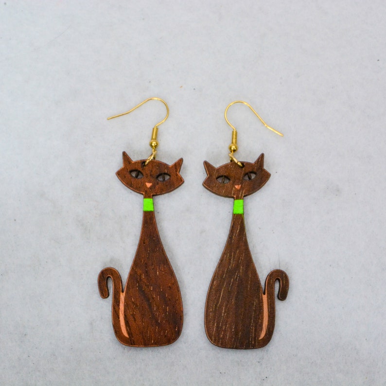 Pode incluir: Um par de brincos de gato de madeira. Os brincos apresentam uma silhueta de gato castanho escuro com uma gola verde e ganchos de orelha dourados. O rosto do gato tem pequenos detalhes laranja. Os brincos s&atilde;o projetados com uma est&eacute;tica moderna de meados do s&eacute;culo.
