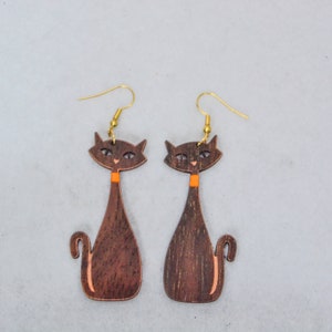 Pode incluir: Um par de brincos de gato de madeira. Os brincos apresentam uma silhueta de gato castanho escuro com uma gola laranja e ganchos de orelha dourados. O rosto do gato tem olhos e nariz pequenos e detalhados.