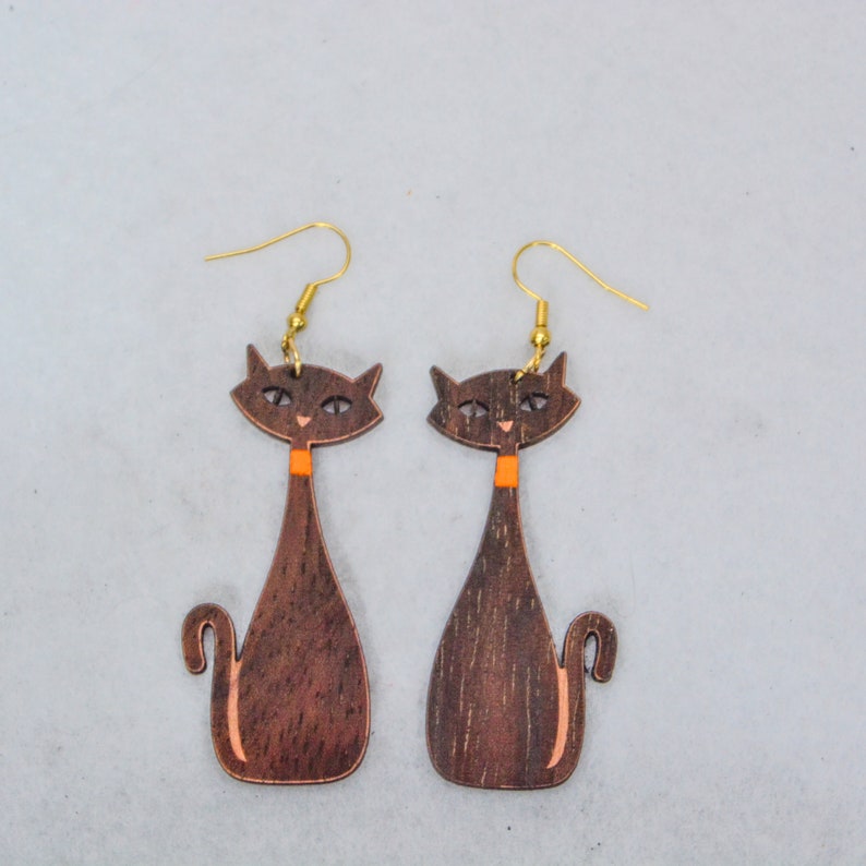 Pode incluir: Um par de brincos de gato de madeira. Os brincos apresentam uma silhueta de gato castanho escuro com uma gola laranja e ganchos de orelha dourados. O rosto do gato tem olhos e nariz pequenos e detalhados.