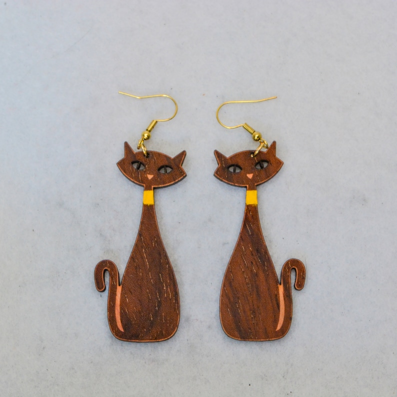 Pode incluir: Um par de brincos de gato de madeira. Os brincos apresentam um design de gato castanho com uma gola amarela e ganchos de orelha dourados. Os olhos e o nariz do gato s&atilde;o pintados de castanho claro. Os brincos t&ecirc;m um estilo moderno de meados do s&eacute;culo.