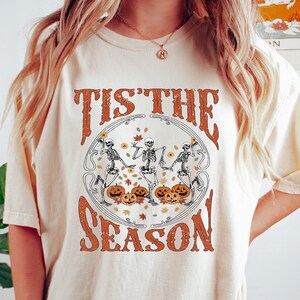 May include: A cream colored t-shirt with a graphic print of three skeletons dancing around pumpkins with the text "Tis the Season".