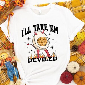 May include: White t-shirt with a graphic of a deviled egg with horns and a tail, standing on fire. The text "I'll take 'em deviled" is above the egg.