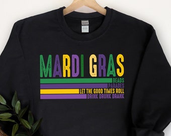 May include: Black sweatshirt with the text "Mardi Gras" in green, yellow, and purple. The text is layered over a series of stripes in the same colors. Below the text is the phrase "Let the good times roll" and "Drink drunk drank" in white.