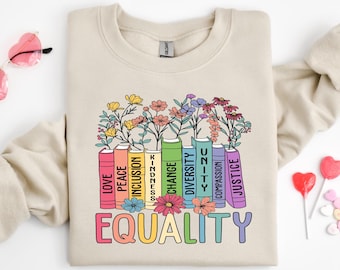 May include: A beige sweatshirt with a rainbow-colored design of books with words like "love", "peace", "inclusion", "kindness", "change", "diversity", "unity", "compassion", and "justice". The words are arranged in a stack of books with flowers growing out of the top. The words "EQUALITY" are printed below the books.