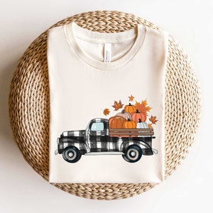 May include: A white t-shirt with a black and white checkered truck carrying pumpkins and fall leaves. The truck has a black and white checkered design and is carrying a variety of pumpkins in orange and brown colors. The truck is surrounded by fall leaves in shades of orange, red, and yellow.