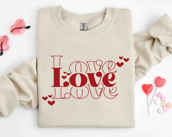 May include: A beige sweatshirt with the word "LOVE" printed in red letters, surrounded by smaller red hearts. The sweatshirt is folded and laid flat on a white surface.