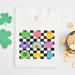 Pode incluir: Camiseta branca com um design xadrez que apresenta v&aacute;rios s&iacute;mbolos do Dia de S&atilde;o Patr&iacute;cio, incluindo um trevo, uma ferradura, um arco-&iacute;ris, um pote de ouro e um cora&ccedil;&atilde;o.