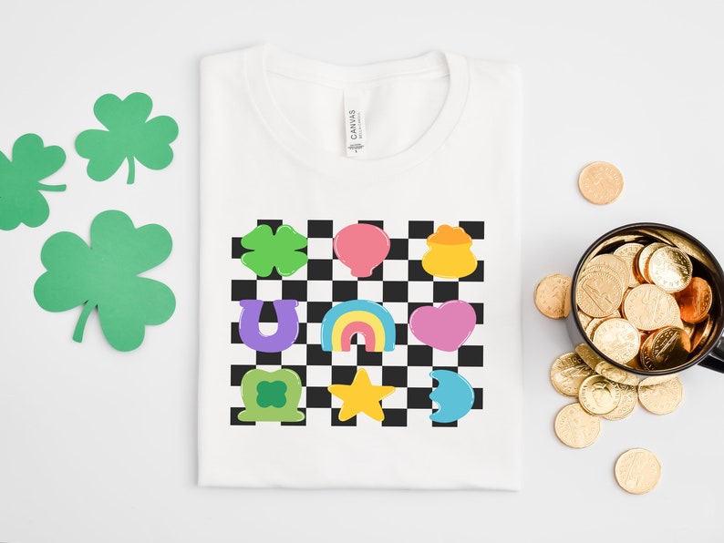 Pode incluir: Camiseta branca com um design xadrez que apresenta v&aacute;rios s&iacute;mbolos do Dia de S&atilde;o Patr&iacute;cio, incluindo um trevo, uma ferradura, um arco-&iacute;ris, um pote de ouro e um cora&ccedil;&atilde;o.