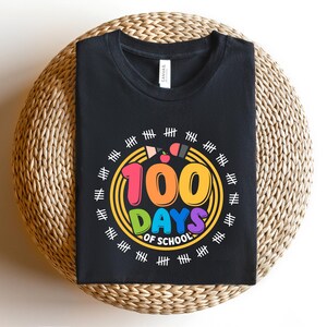 May include: Black t-shirt with a colorful graphic celebrating 100 days of school. The design features the text "100 DAYS OF SCHOOL" in vibrant colors, surrounded by a circle with pencils and tally marks. The shirt is displayed on a woven surface.