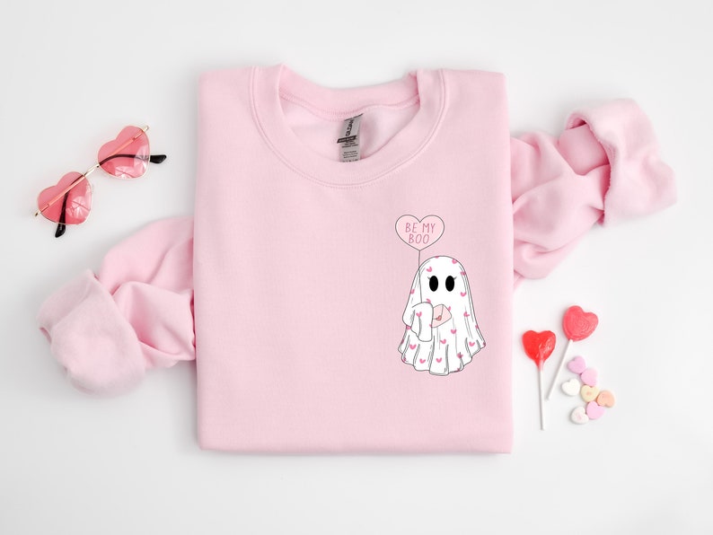 Op de afbeelding: Roze sweatshirt met een witte geest die een hartvormige ballon vasthoudt met de tekst "Be My Boo".