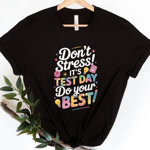 May include: Black t-shirt with the text "Don't Stress! It's Test Day Do your BEST!" in a colorful design. The design includes a smiley face, light bulbs, a book, and a pencil. The shirt is hanging on a wooden hanger.