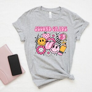May include: Gray t-shirt with the text "Second Grade" in pink letters. The shirt features a graphic design of a pink tongue, smiley faces, a flower, a globe, a pencil, and a checkered pattern.