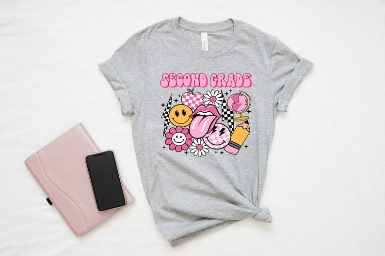 May include: Gray t-shirt with the text "Second Grade" in pink letters. The shirt features a graphic design of a pink tongue, smiley faces, a flower, a globe, a pencil, and a checkered pattern.