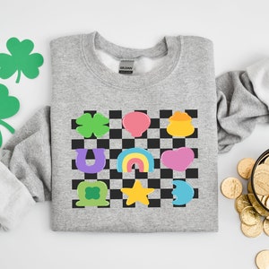 Pode incluir: Uma camisa de moletom cinza com um padr&atilde;o xadrez que apresenta v&aacute;rios amuletos da sorte, incluindo um trevo de quatro folhas, uma ferradura, um arco-&iacute;ris, um pote de ouro e uma estrela. Os amuletos s&atilde;o de diferentes cores, incluindo verde, roxo, amarelo, rosa e azul.