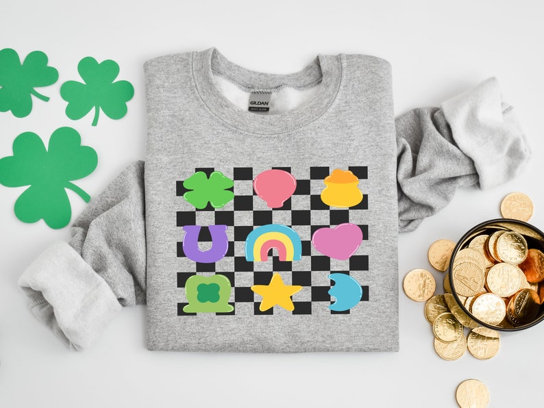 Pode incluir: Uma camisa de moletom cinza com um padr&atilde;o xadrez que apresenta v&aacute;rios amuletos da sorte, incluindo um trevo de quatro folhas, uma ferradura, um arco-&iacute;ris, um pote de ouro e uma estrela. Os amuletos s&atilde;o de diferentes cores, incluindo verde, roxo, amarelo, rosa e azul.