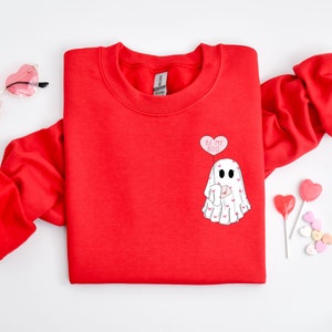 Op de afbeelding: Rood sweatshirt met een witte geestenprint en de tekst "Be My Boo" in een roze hart. De geest houdt een brief met harten erop.