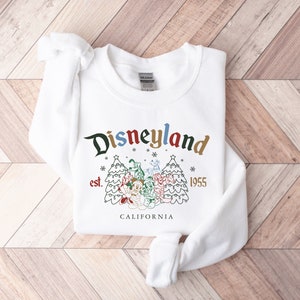 May include: White sweatshirt with a Christmas-themed Disneyland graphic. The graphic features a stylized Disneyland logo with the text "Disneyland est. 1955 CALIFORNIA" and a cartoon image of Mickey Mouse and friends standing in front of two Christmas trees.