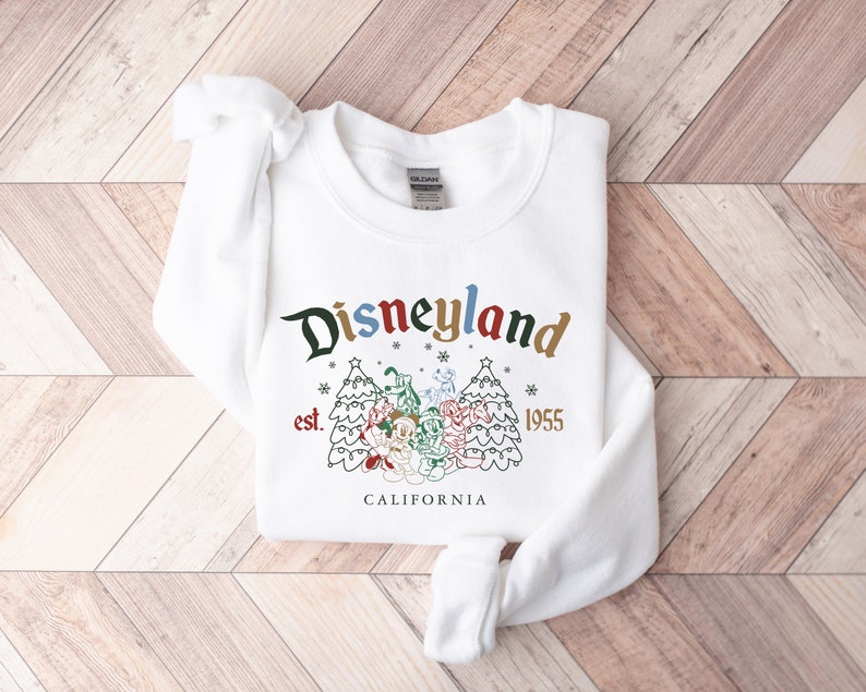 May include: White sweatshirt with a Christmas-themed Disneyland graphic. The graphic features a stylized Disneyland logo with the text "Disneyland est. 1955 CALIFORNIA" and a cartoon image of Mickey Mouse and friends standing in front of two Christmas trees.