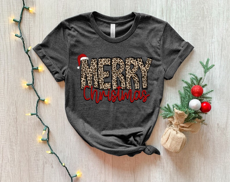 May include: A dark gray t-shirt with a leopard print "Merry Christmas" design. The word "Merry" is in a leopard print with a red Santa hat on top of the "M". The word "Christmas" is in red.