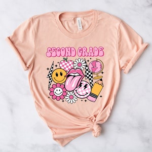 May include: A peach-colored t-shirt with the text "Second Grade" in pink letters. The shirt features a variety of pink and yellow graphic designs, including a smiley face, a flower, a checkered pattern, a tongue, a pencil, and a globe.